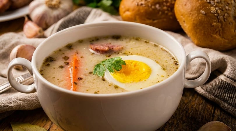 Creamy soup with a hard-boiled egg, sausage, carrot, and parsley in a white bowl.