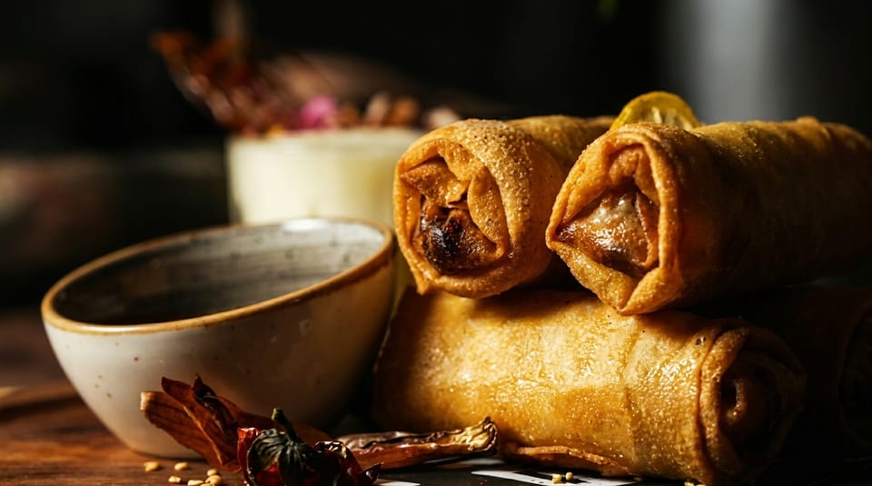 Golden-brown spring rolls with a dipping sauce bowl and dried chilies on a dark wooden surface.