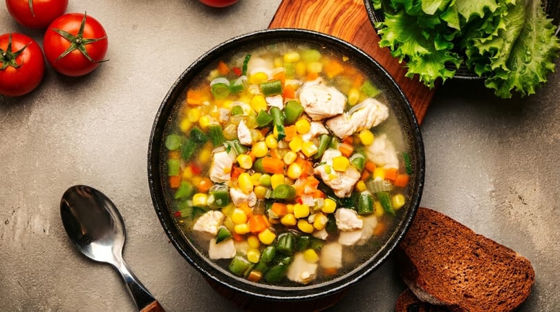 Hearty chicken and vegetable soup with corn, carrots, and green beans, served with bread and tomatoes.