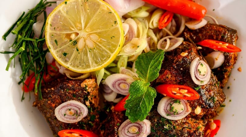 Close-up of spicy fish with lemon, red chilies, sliced onions, and fresh herbs.